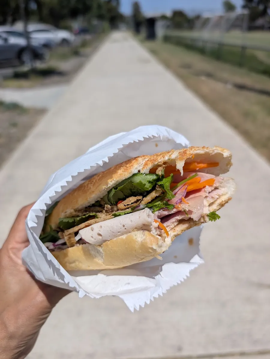 A Glengala banh mi wrapped in white paper, filled with ham, pork, fresh herbs, carrot and cucumber.