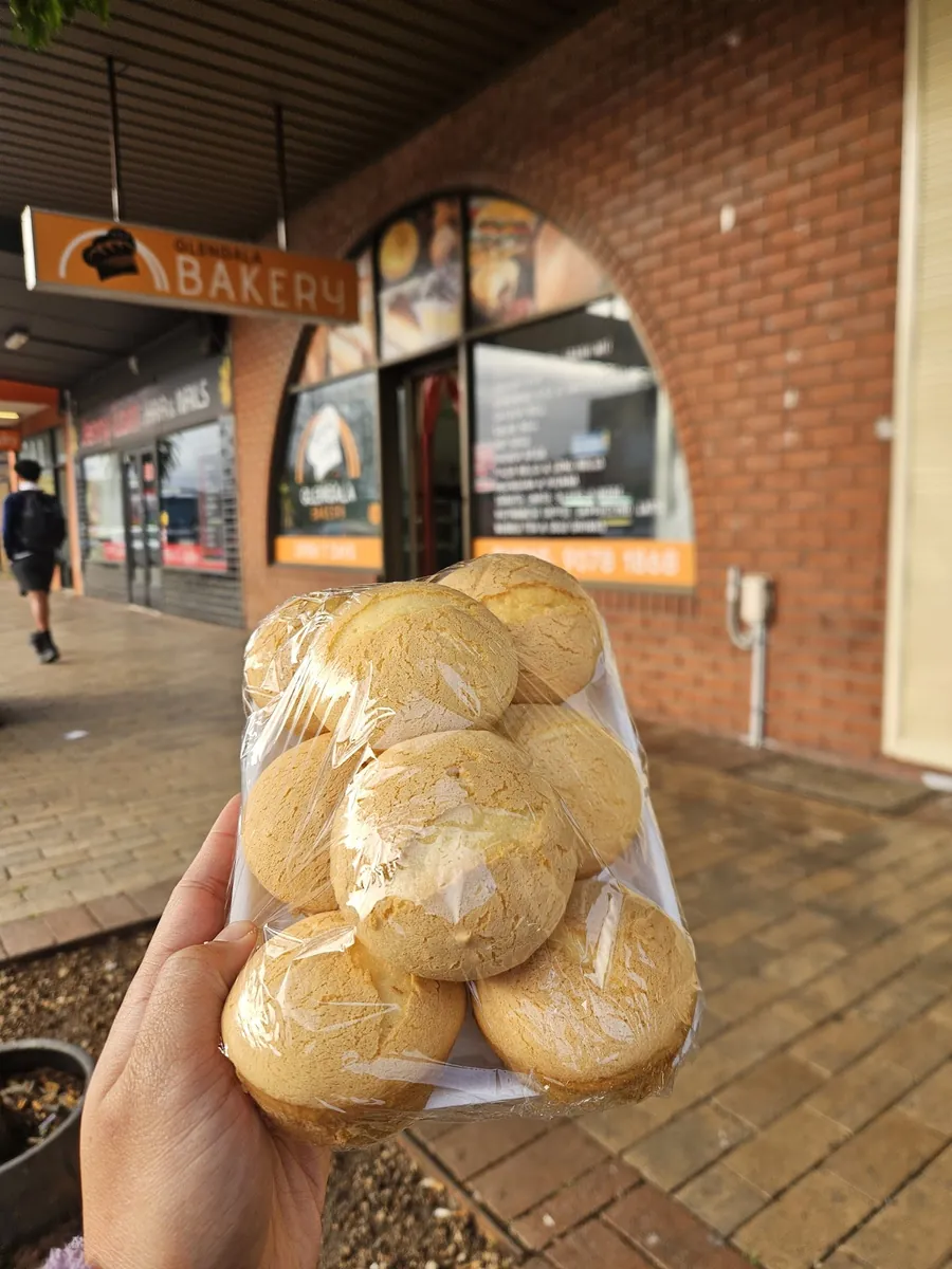 Glengala Bakery shopfront in Glengala Village: an orange sign and a brick arch window, with a bag of fresh bread rolls held up in front.